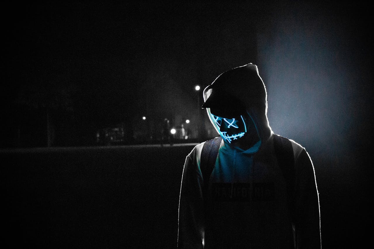 our-services-1 A mysterious figure wearing a LED mask and hoodie stands illuminated in a dark, foggy street at night.